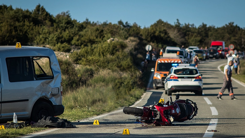 A traffic accident involving a motorcycle and a van on a sunny, two-lane road in a relatively rural or open area.