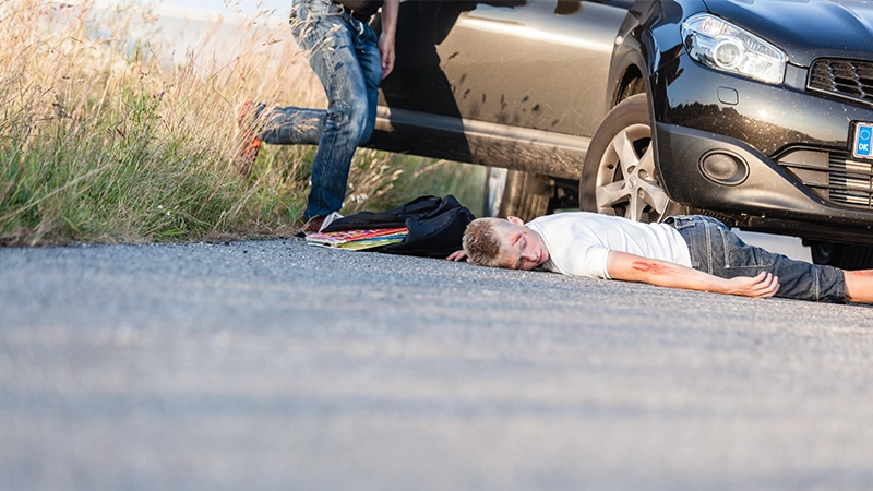 A boy hit by a vehicle, lying on the road wounded and the car owner running towards him for help.