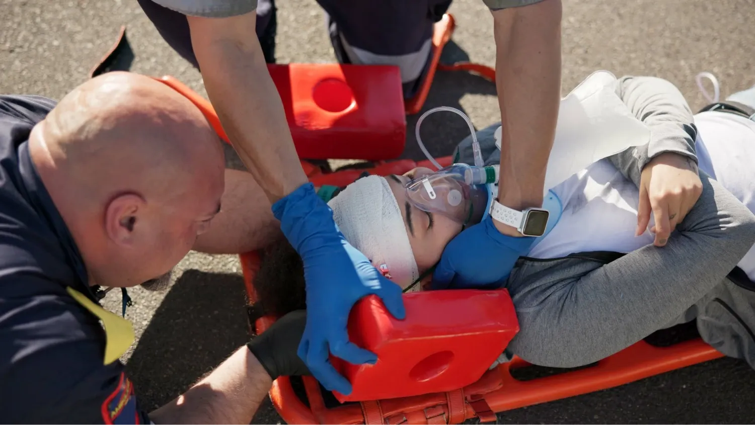 The injured person has a head injury wearing an oxygen mask, she is lying down while receiving care, with one paramedic.