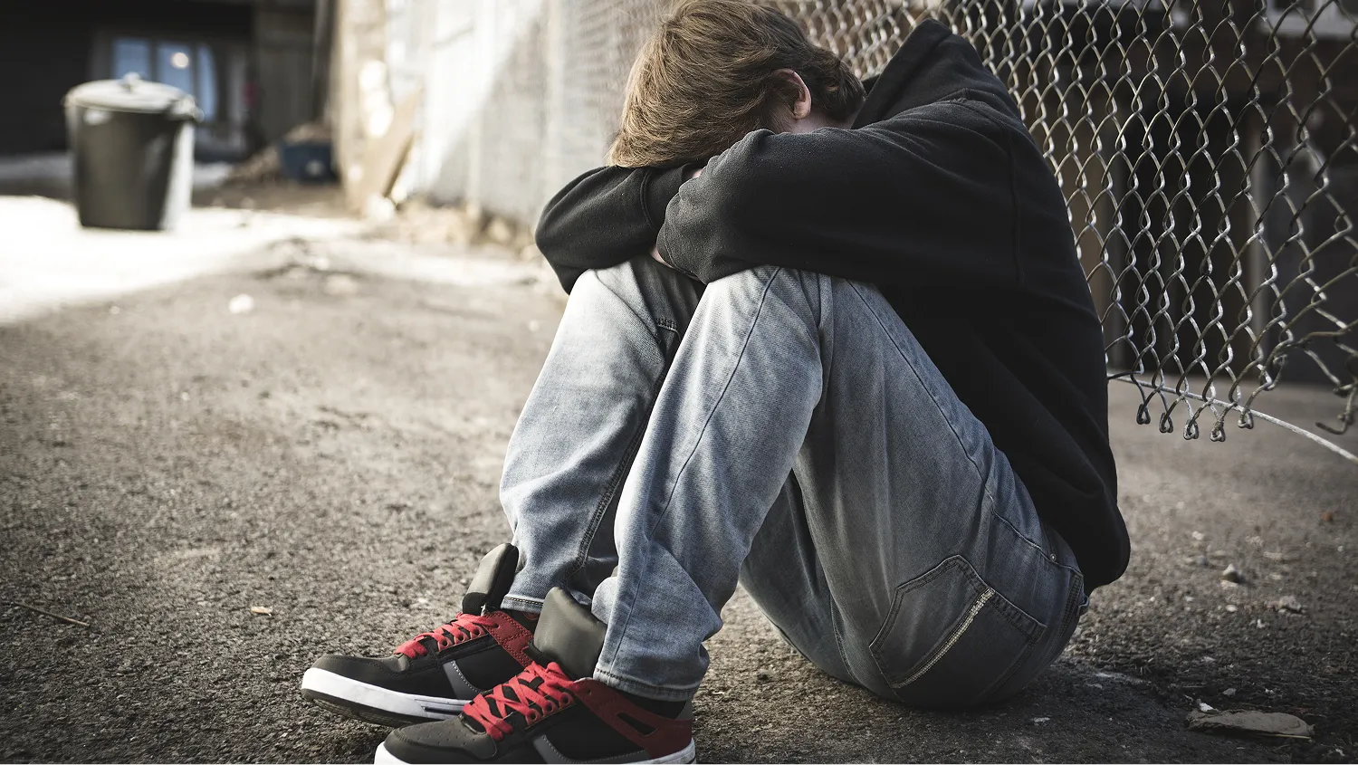 A young person curled up and sitting on the ground in an alleyway, with their face hidden, he is wearing a black hoodie and jeans.