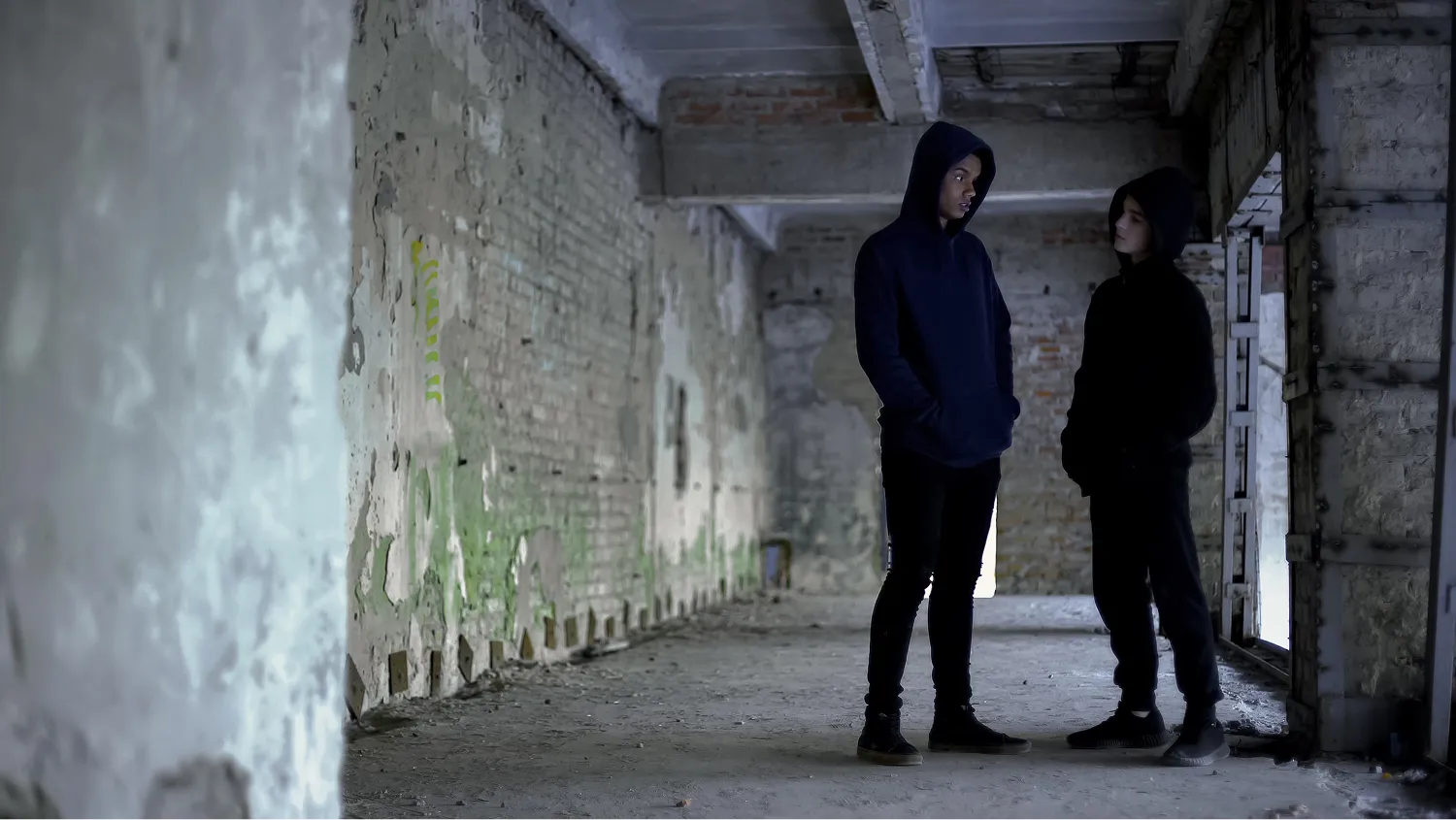 Two male youths stand facing each other in a dimly lit, dilapidated building, wearing dark hoodies and pant discussing something.