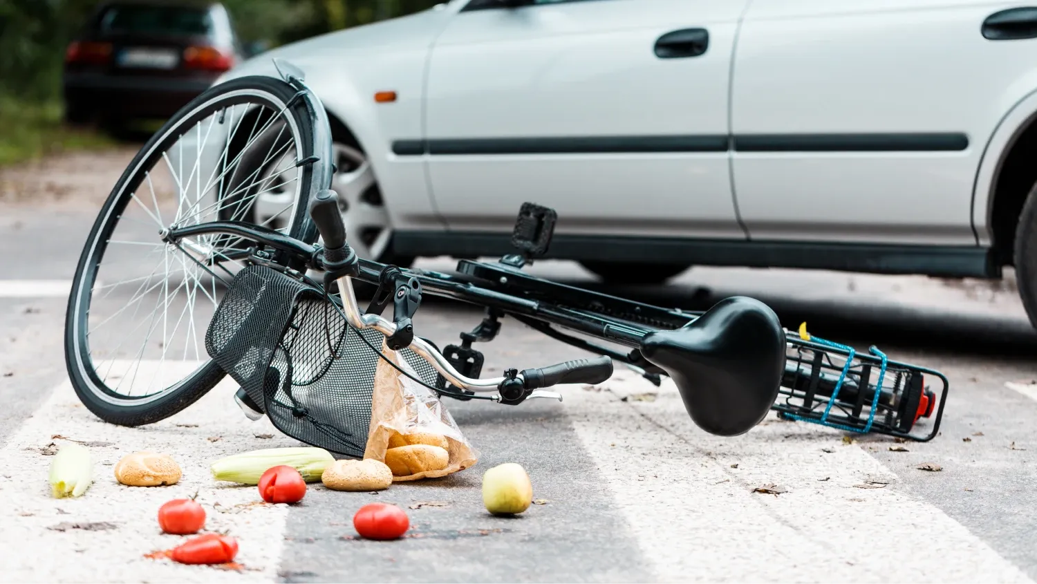 a crumpled bicycle and a silver car after a bicycle accident, and a paper bag with groceries, including tomatoes and other produce, has spilled into the road.
