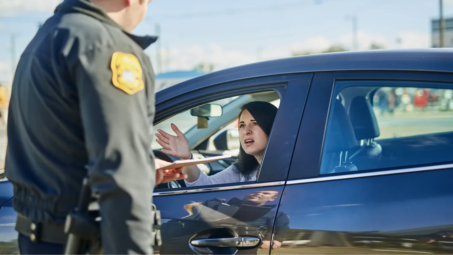 A police office speaking to a young woman who is in the driver's seat of a dark-colored car.