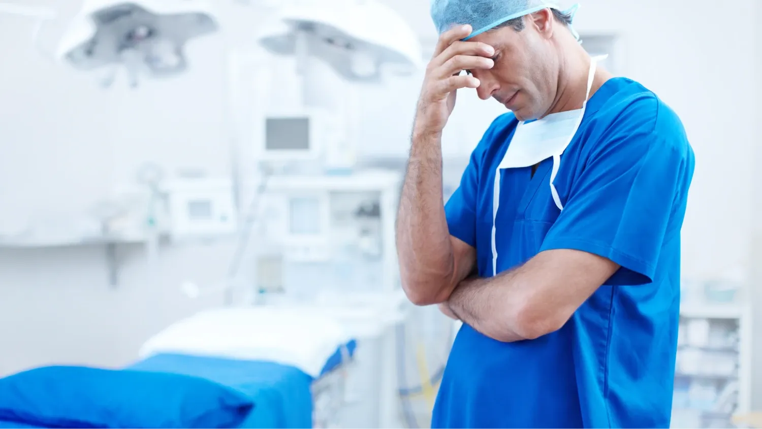 a frustrated male surgeon in blue scrubs, a surgical cap, and a mask pulled down around his neck, and standing in operating room, with his hand on his forehead and his eyes close.