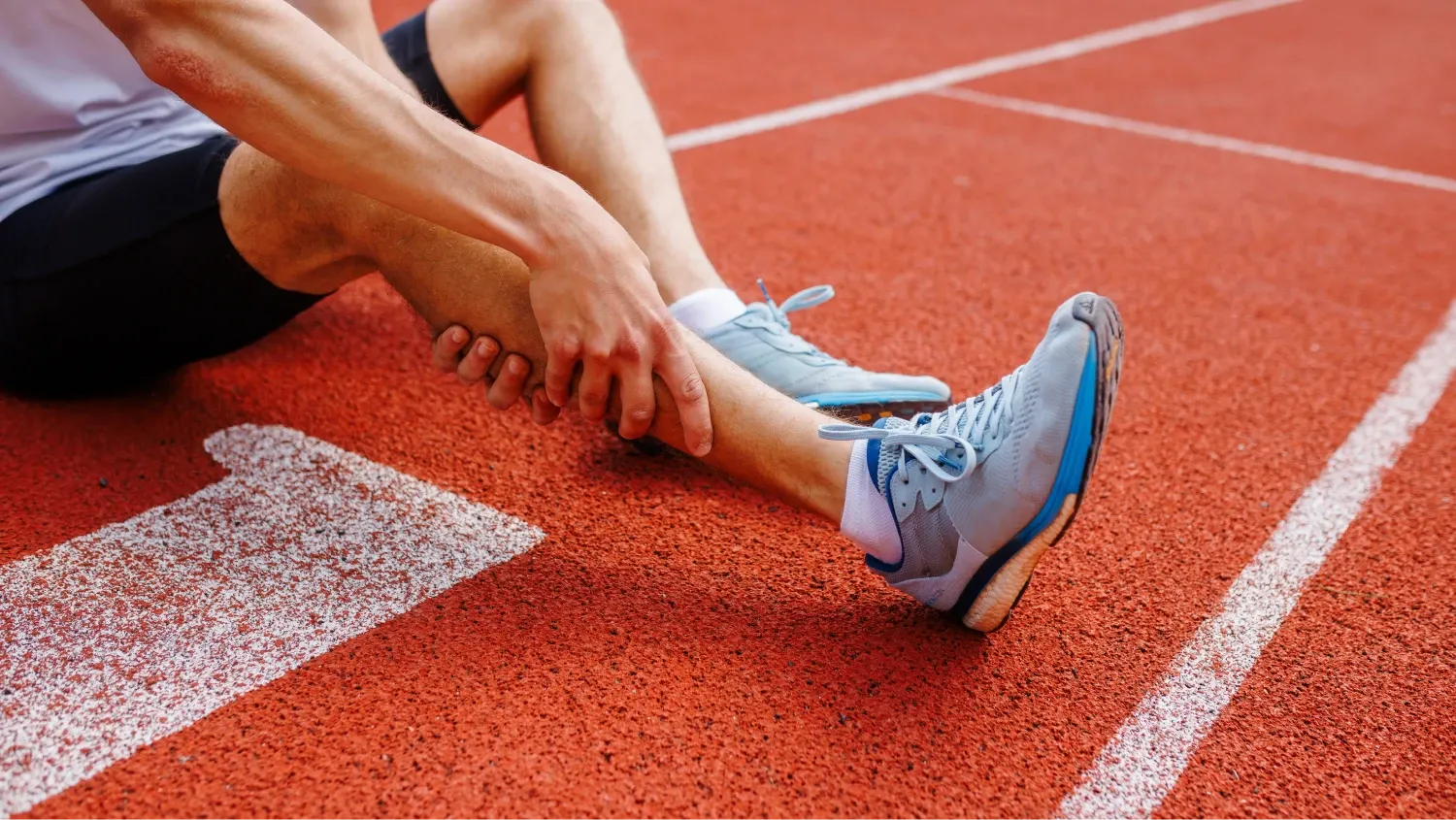 A male athlete sitting on a red running track. He is holding his lower leg and ankle with his hand, a gesture commonly associated with an injury.