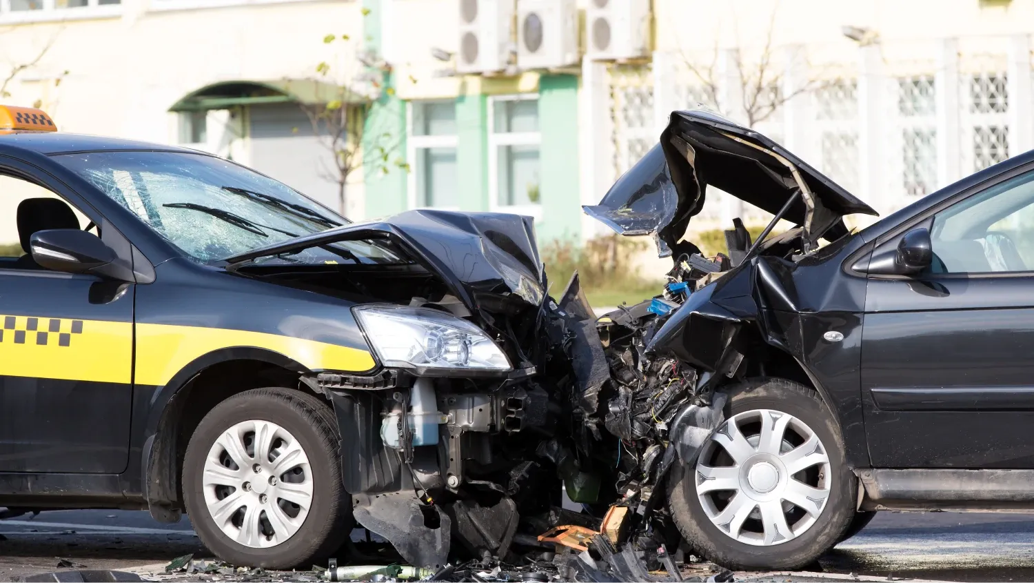 A head-on collision between two cars. A taxi cab, identifiable by its yellow-and-black checkered stripe and roof light, has its front end completely crushed.