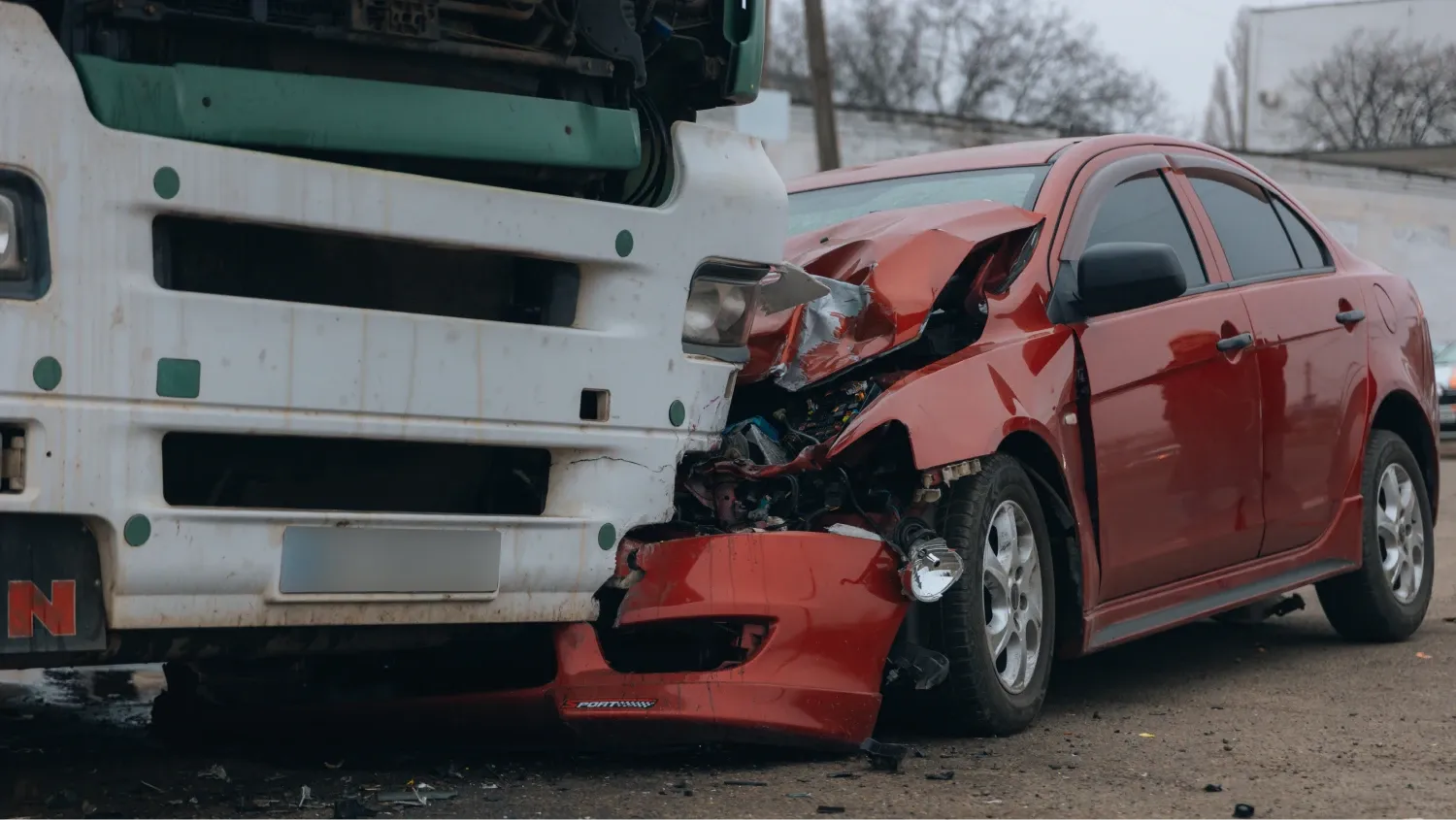A serious vehicle collision between a large truck and a red sedan, and the front of the sedan has been completely crushed and is wedged under the front bumper of the truck.