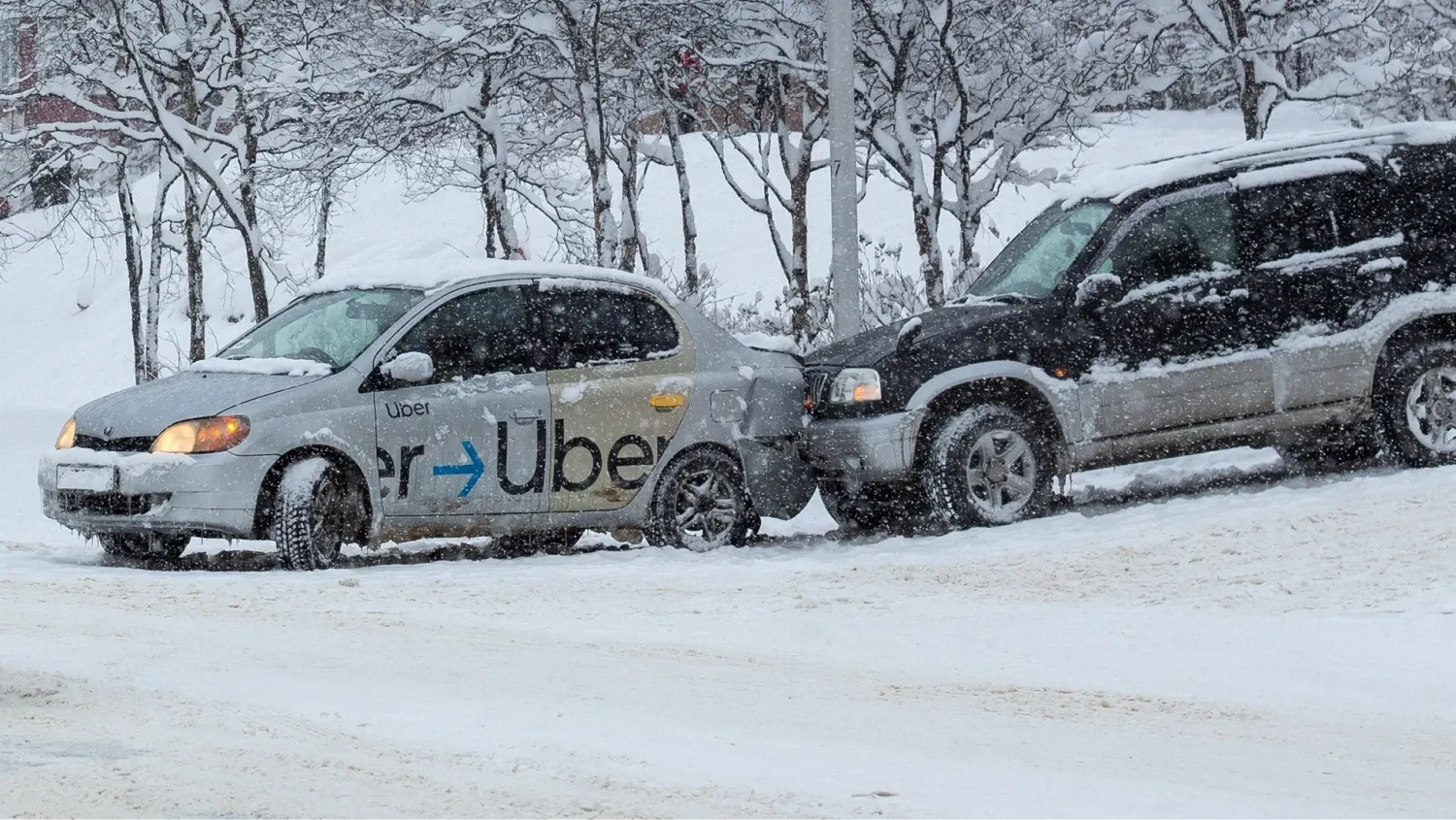 A silver sedan with a "Uber" decal on its side has been rear-ended by a dark SUV. Both vehicles are covered in snow, and the street and trees in the background are also blanketed in white.
